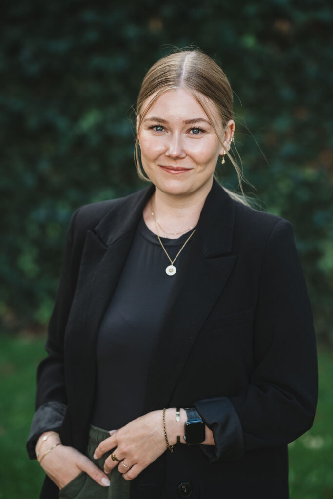 Marketingassistent Ninna står smilende foran en grøn busk.